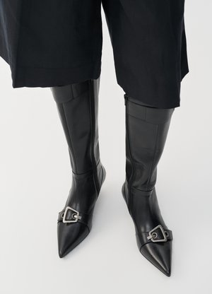 Bottes noires en cuir montantes avec un bout pointu et un accent de boucle en argent. Présentent une fermeture éclair sur le côté intérieur et une texture lisse.