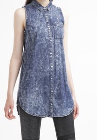 Camicia senza maniche in denim, azzurro chiaro con lavaggio sbiadito, chiusura frontale con bottoni, colletto e taschino singolo sul petto. Orlo curvo nella parte inferiore.