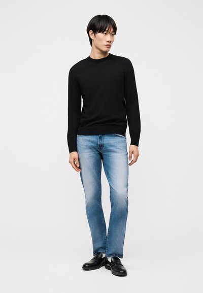 Νεαρός άντρας που στέκεται φορώντας μαύρο πουλόβερ με μακριά μανίκια, ανοιχτόχρωμα ξεθωριασμένα τζιν και μαύρα δερμάτινα παπούτσια, μπροστά σε απλό ανοιχτό φόντο.