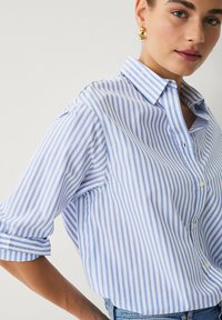 Camisa abotonada de rayas azul claro y blanca, hecha de algodón, con cuello puntiagudo y mangas arremangadas.