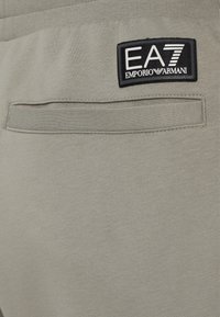 Svetlosivé tepláky s elastickým pásom, obsahujúce zadné vrecko a čiernu nášivku loga EA7 Emporio Armani. Mäkkučká, textúrovaná tkanina.