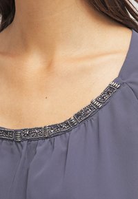 Blusa grigia con scollatura rotonda decorata con abbellimenti in perline nei toni argento e scuri, realizzata in un tessuto morbido e fluido.
