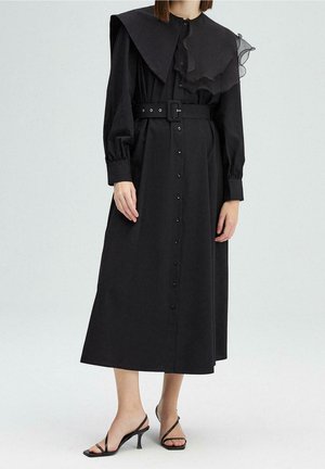 Kobieta ubrana w długą czarną sukienkę na guziki z szerokim kołnierzem, paskiem w talii, długimi rękawami oraz czarne sandały na obcasie z paskami.