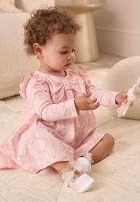 Vestito rosa chiaro con volant e dettaglio a fiocco, decorato con un motivo di fiocchi. Il bambino tiene in mano un piccolo coniglio di crochet. Scarpe morbide bianche con accenti.