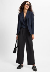 Marineblauer Nadelstreifenblazer über einem schwarzen Oberteil, kombiniert mit weiten schwarzen Hosen. Die Accessoires umfassen übergroße Sonnenbrillen und eine schwarze Handtasche.