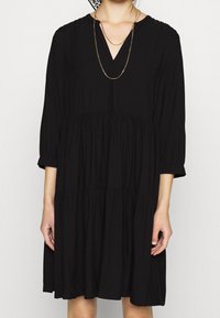 Schwarzes Kleid aus weichem Stoff, mit V-Ausschnitt, Dreiviertelärmeln und einem gestuften, gerafften Rockdesign. Akzentuierte dünne Kettenhalskette.