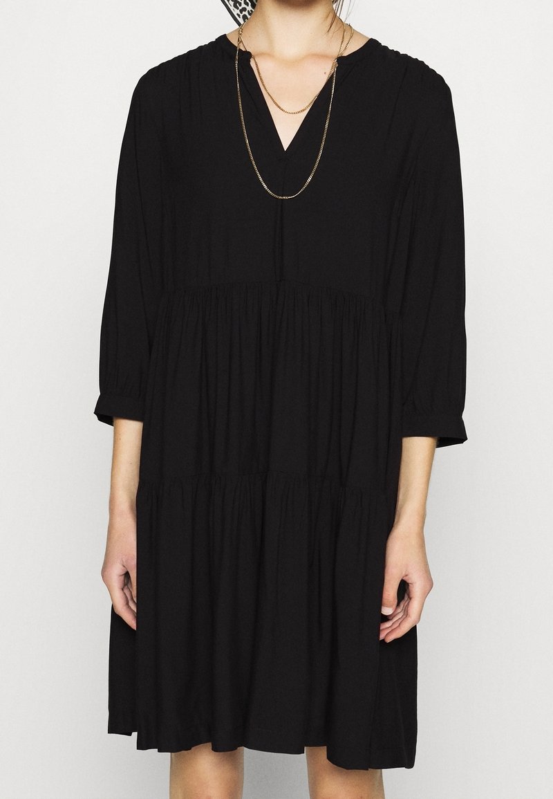Schwarzes Kleid aus weichem Stoff, mit V-Ausschnitt, Dreiviertelärmeln und einem gestuften, gerafften Rockdesign. Akzentuierte dünne Kettenhalskette.