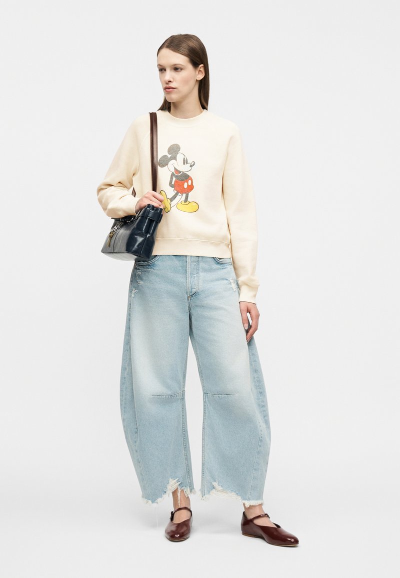 Femme portant un pull crème avec un imprimé Mickey Mouse, un jean bleu clair déchiré à larges jambes, des ballerines bordeaux, et tenant un sac à main noir sur l'épaule.