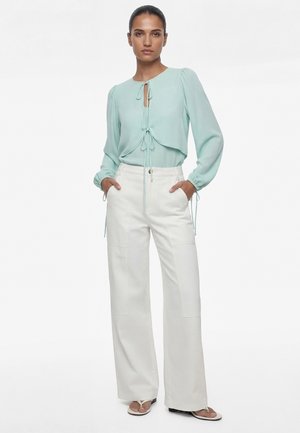 Kvinde iført en lyseblå bluse med lange ærmer og bindebånd samt hvide bukser med vidde, stående med hænderne i lommerne foran en hvid baggrund.