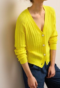 Cardigan giallo a maglia con scollo a V profondo, maniche lunghe e bottoni a forma di fiore in argento. Tessuto texturizzato con vestibilità ampia, abbinato a jeans blu.