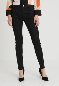 Femme portant un pantalon noir slim pailleté et des chaussures à talons hauts noires, debout devant un fond clair uni.