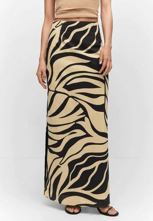 Femme portant une jupe maxi beige taille haute avec de grands motifs abstraits noirs de feuilles et des sandales noires à bout ouvert.