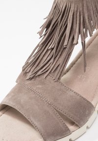 Gros plan d'une sandale en daim taupe avec un détail de franges superposées et de larges brides cousues sur une semelle rembourrée beige.