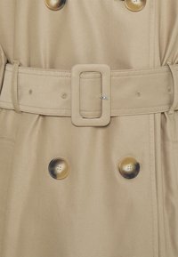 Cappotto beige con design doppiopetto, grandi bottoni rotondi, una cintura coordinata con fibbia quadrata e una texture del tessuto liscia.