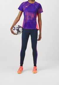Camisa deportiva de manga corta con estampado morado y rosa, leggings azul marino con acentos laterales, y zapatillas de fútbol de color naranja brillante, sosteniendo un balón de fútbol.