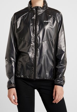 Personne portant une veste noire brillante zippée avec des poignets élastiques et un petit logo blanc sur la poitrine, associée à un pantalon noir.