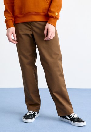 Person iført brune bukser, sorte og hvide sneakers og en orange sweatshirt, der står på en lyseblå gulv mod en ensfarvet hvid baggrund.