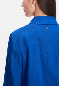 Blue linen shirt with a collar, visible stitching, and a small metal logo at the back. The fabric has a slight texture.