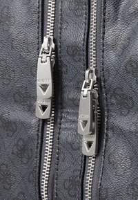 Matériau synthétique noir texturé avec des zips argentés. Les tirettes des zips présentent un design triangulaire et un branding. Gros plan sur les zips en détail.
