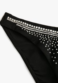 Svart bikinitrosa med en sl�ät textur, prydd med silverfärgade nitar och kvadratiska detaljer längs midjebandet, vilket skapar ett dekorativt mönster.