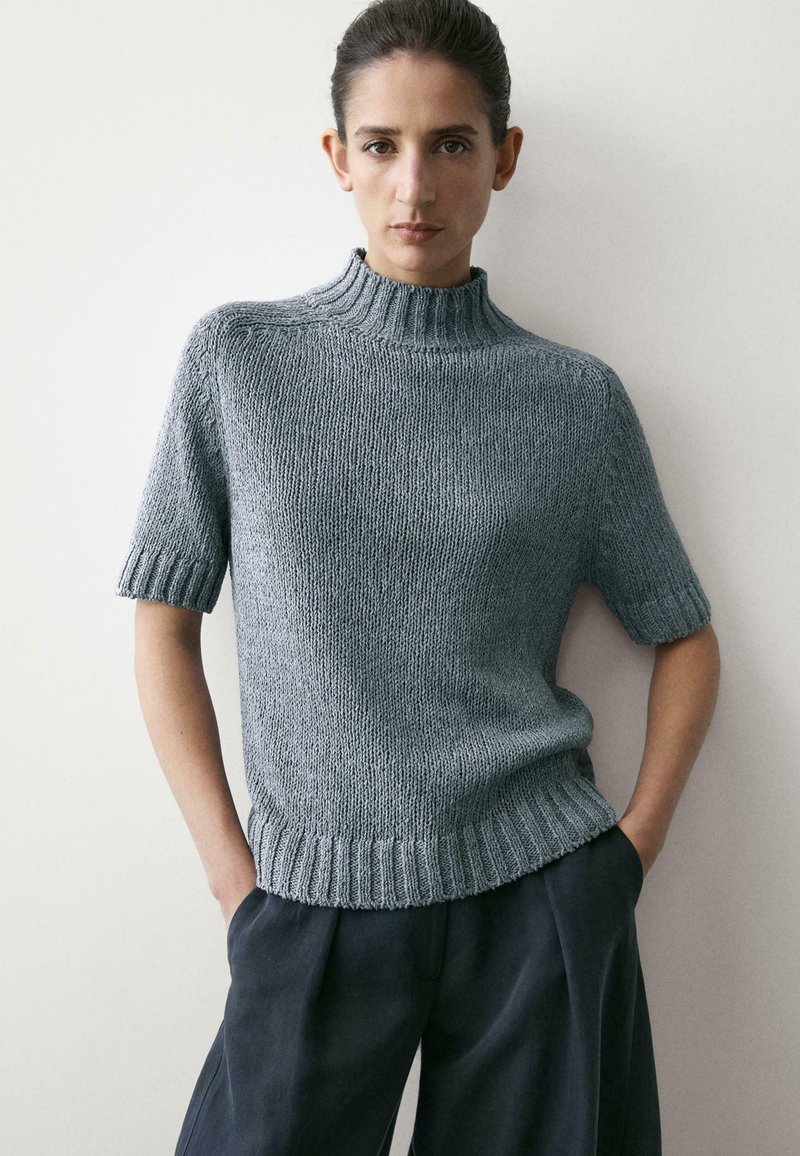 Kvinde med mørkt hår iført en grå kortærmet striktrøje og mørke bukser, stående foran en lys, ensfarvet væg med hænderne i lommerne.