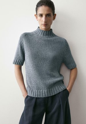 Vrouw met donker haar in een grijze korte mouw gebreide trui en donkere broek, staand tegen een lichte effen muur met handen in de zakken.