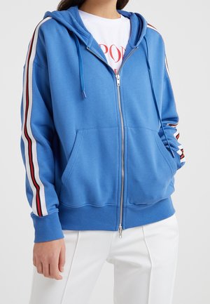 Blauer Kapuzenpullover mit Reißverschluss und roten, weißen sowie schwarzen Streifen an den Ärmeln, getragen über einem weißen Shirt und weißen Hosen, Hände in den Taschen.