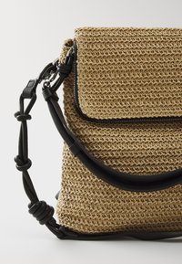 Vevd stråveske med sort lærreim, med en klaff som kan foldes over og glidelåsdetalj. Naturlig beige farge med en teksturert overflate.