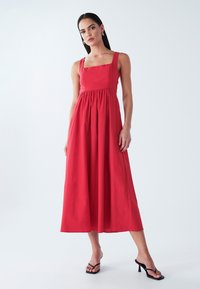 Rotes, ärmelloses Maxikleid mit quadratischem Ausschnitt, geraffter Taille und fließendem Rock. Glatter Stoff; Modell trägt schwarze Flip-Flops.