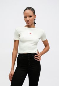 Bela obrezana majica z kratkimi rokavi s teksturiranim vzorcem in rdečo vezenino srca na prsih, kombinirana z visokim pasom črnih kavbojk.