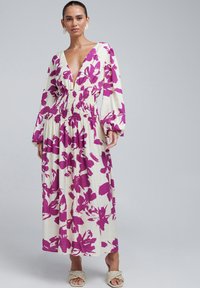 Robe maxi florale en crème avec des motifs de fleurs violettes audacieux. Elle présente un décolleté en V profond, une taille smockée et de longues manches bouffantes. Tissu texturé.
