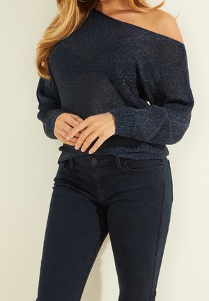 Navy off-the-shoulder sweater with a textured, shimmery fabric. Features long sleeves, ribbed cuffs, and a relaxed fit, paired with dark jeans.