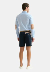 Camicia button-up azzurro chiaro con toppe sui gomiti, pantaloni corti blu navy, cintura beige e sneakers bianche. Vista posteriore, che mostra i dettagli della camicia blu e dei pantaloni.
