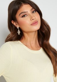 Maglione a coste giallo chiaro con scollatura a girocollo, tessuto testurizzato, indossato da un modello che porta orecchini a goccia floreali, mettendo in evidenza dettagli delicati.