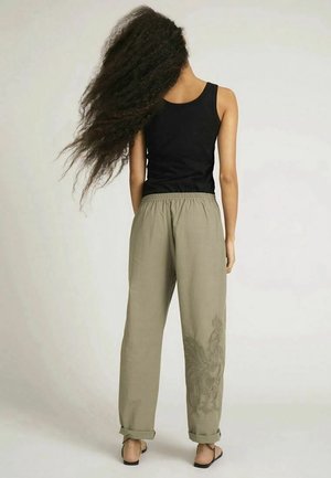 Pantalons légers couleur kaki avec une coupe décontractée et une taille élastique, présentant un motif floral subtil sur la jambe gauche. Associés à un haut noir à bretelles.