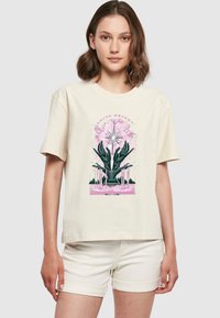 Beige t-skjorte med kort erme med et fargerikt grafisk design av hender og blomstrende elementer med teksten "SKINN KLART". Myk bomullsmateriale.
