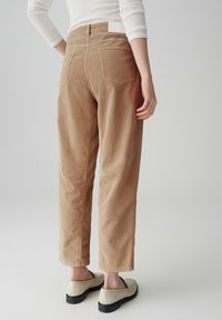 Pantalons en velours côtelé marron avec une coupe décontractée, dotés de poches arrière et d'une étiquette blanche au niveau de la taille. Portés avec des chaussures à enfiler claires.