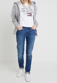 Person iført en grå hættetrøje, hvid grafisk t-shirt med tekst, blå jeans og hvide sneakers, står med hænderne i lommerne mod en hvid baggrund.