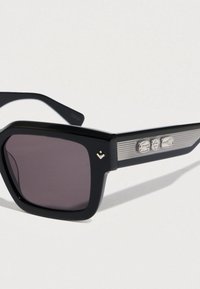 Mustat aurinkolasit, joissa on suorakulmainen kehys, tummat linssit ja hopeiset raidalliset yksityiskohdat varsissa, joissa on kolme pientä koriste-elementtiä.