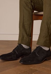 Zwarte suède veterschoenen met een ronde neus, gecombineerd met lichtgrijze sokken. Olijfgroene gestructureerde broek, zittend op een houten stoel.