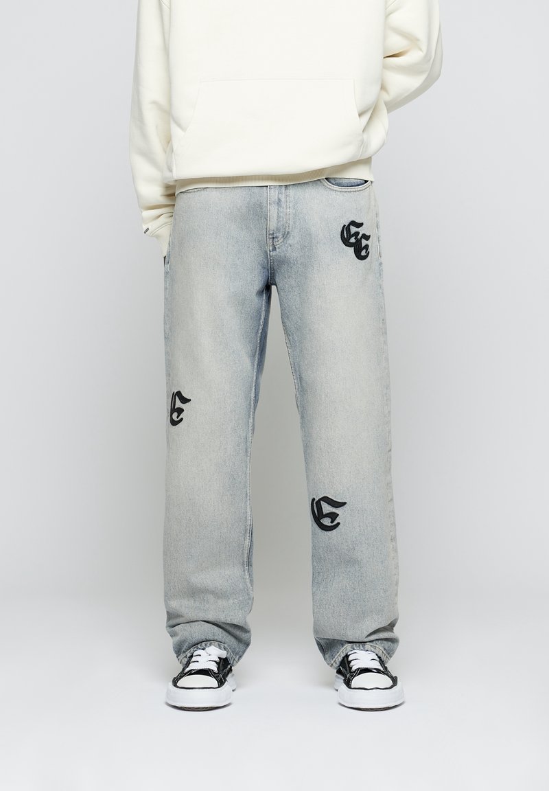 Jeans de mezclilla azul claro con un ajuste holgado. Presentan logotipos bordados en negro en el frente, combinados con una sudadera blanca con capucha y zapatillas negras.