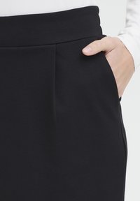 Ruka v kapse černých kalhot s vysokým pasem nošených s bílým dlouhým rukávem na světlém pozadí.