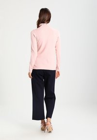 Pull à col montant rose clair en tissu doux, doté de manches longues et d'une coupe ajustée, associé à un pantalon large bleu marine et des talons beiges.