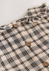 Beige plaid shirt with blue and black lines, featuring a ruffled collar and three wooden buttons along the front. Soft fabric texture.