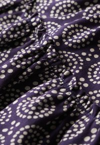 Lila Stoff mit weißem Tupfenmuster, versehen mit gerafften Rüschen und strukturierten Nähten, Nahaufnahme des Materials.