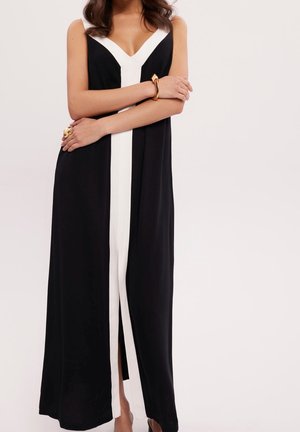 Vrouw draagt een mouwloze zwarte maxi-jurk met een witte V-hals en verticale streep, geaccentueerd met een gouden armband en ring.