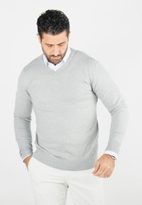 Pull gris en V en matériau doux avec des manches longues, des poignets et un ourlet côtelés, porté par-dessus une chemise blanche à col, associé avec un pantalon clair.