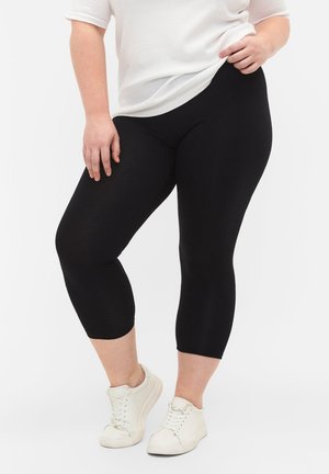 Oseba, oblečena v črne kratke legice, bele superge in belo teksturirano kratko majico z rokavi, rahlo dviguje zgornji rob majice.