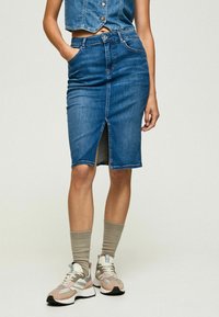 Kvinna som bär en blå denimpennkjol med en framklippa, i kombination med beige strumpor och flerfärgade sneakers.