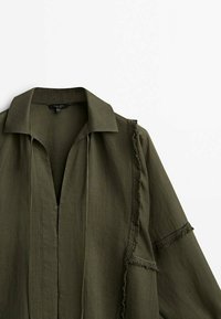 Olivengrøn skjorte med krave og flagrende kanter langs fronten og ærmerne. Lavet af et let, tekstureret stof.